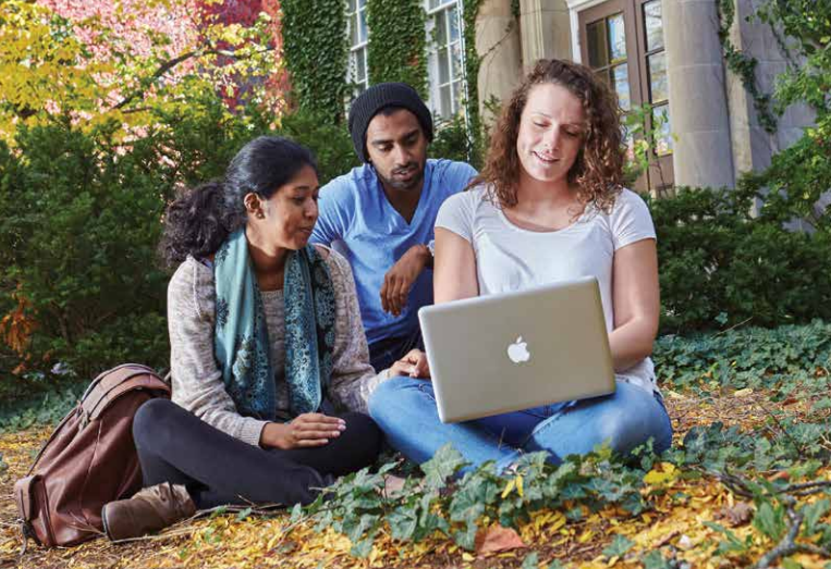 3 students sit outside on campus with a laptop