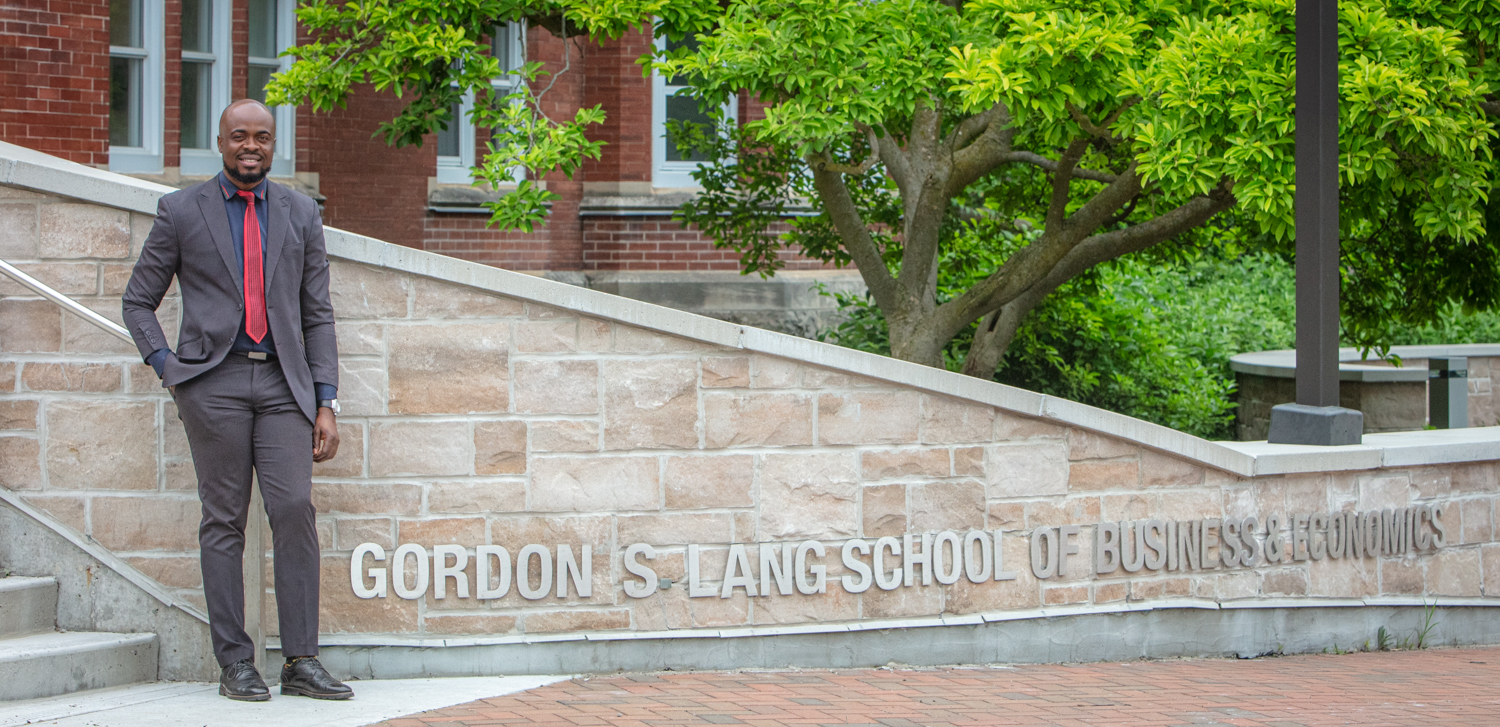 Toochukwu Urama standing in front of a Gordon S Lang School of Business & Economics sign